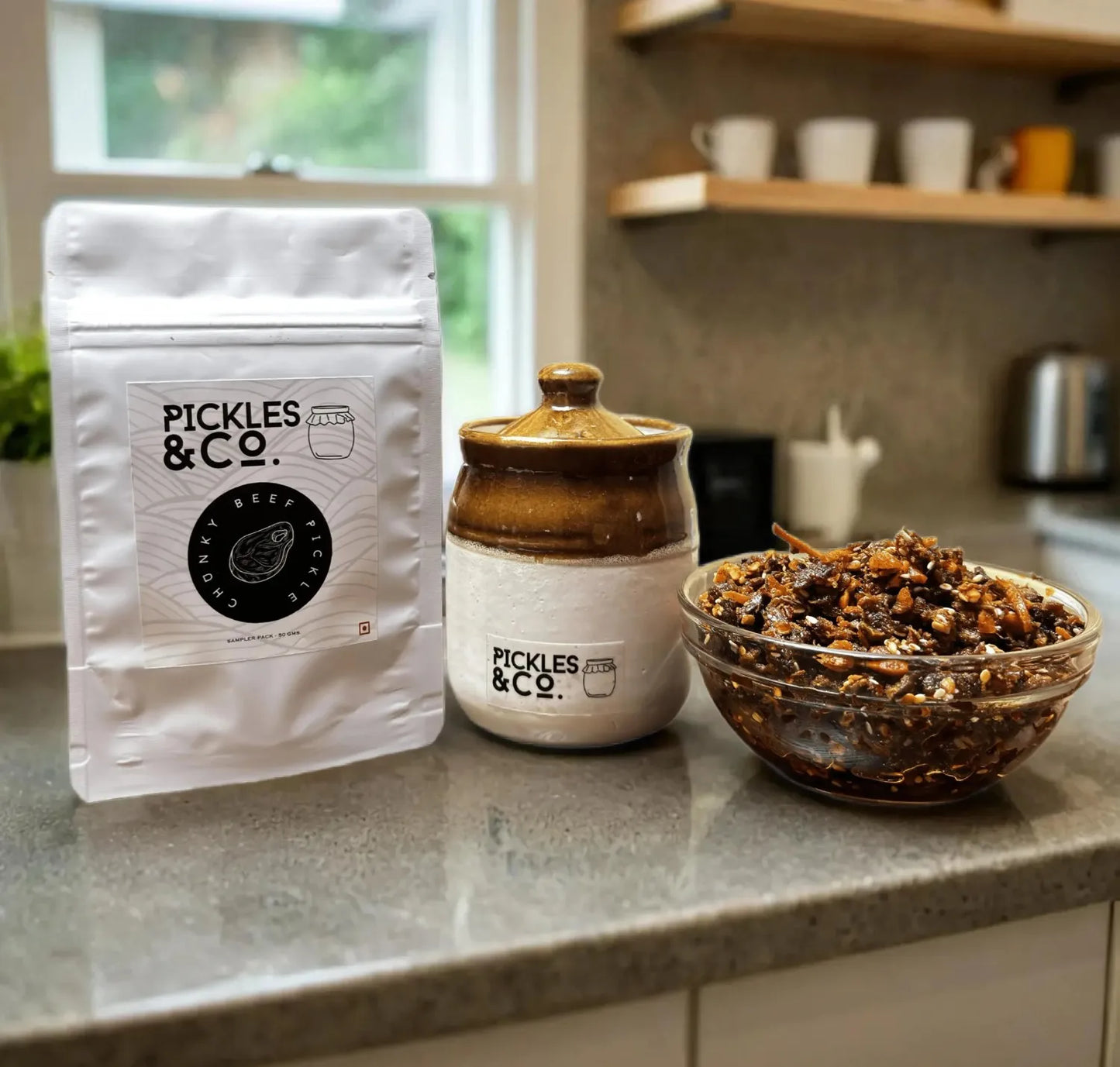 A jar of beef pickle and its packaging on a kitchen counter, with the brand 'PICKLES & CO' visible on the label. A bowl of the pickle is also visible in the foreground.